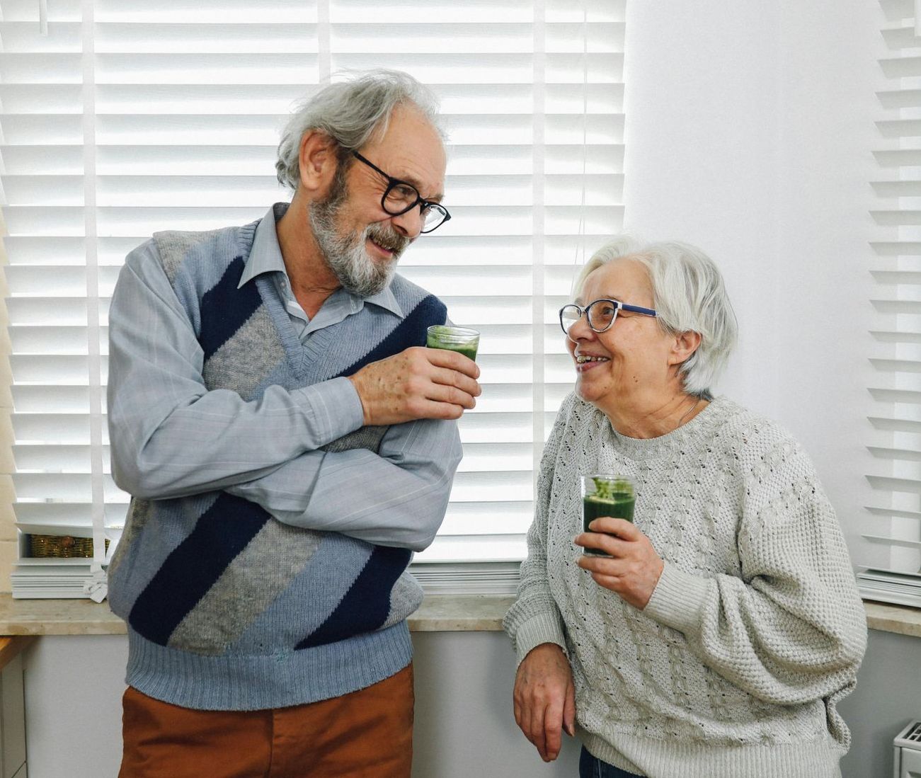 An elderly couple is standing next to each other in front of a window.