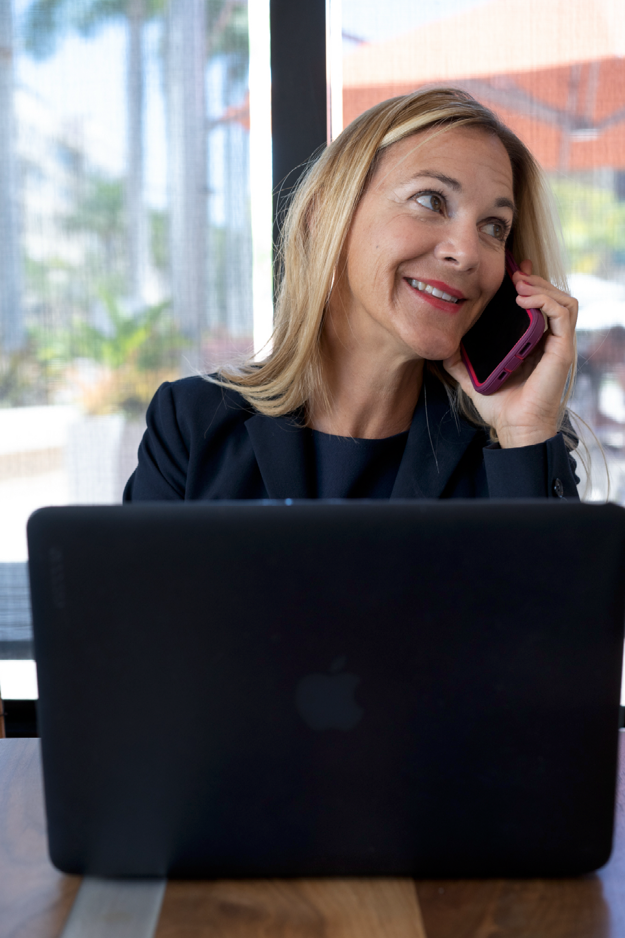 Woman in blazer smiling while talking on a phone, seated at a table with a laptop.
