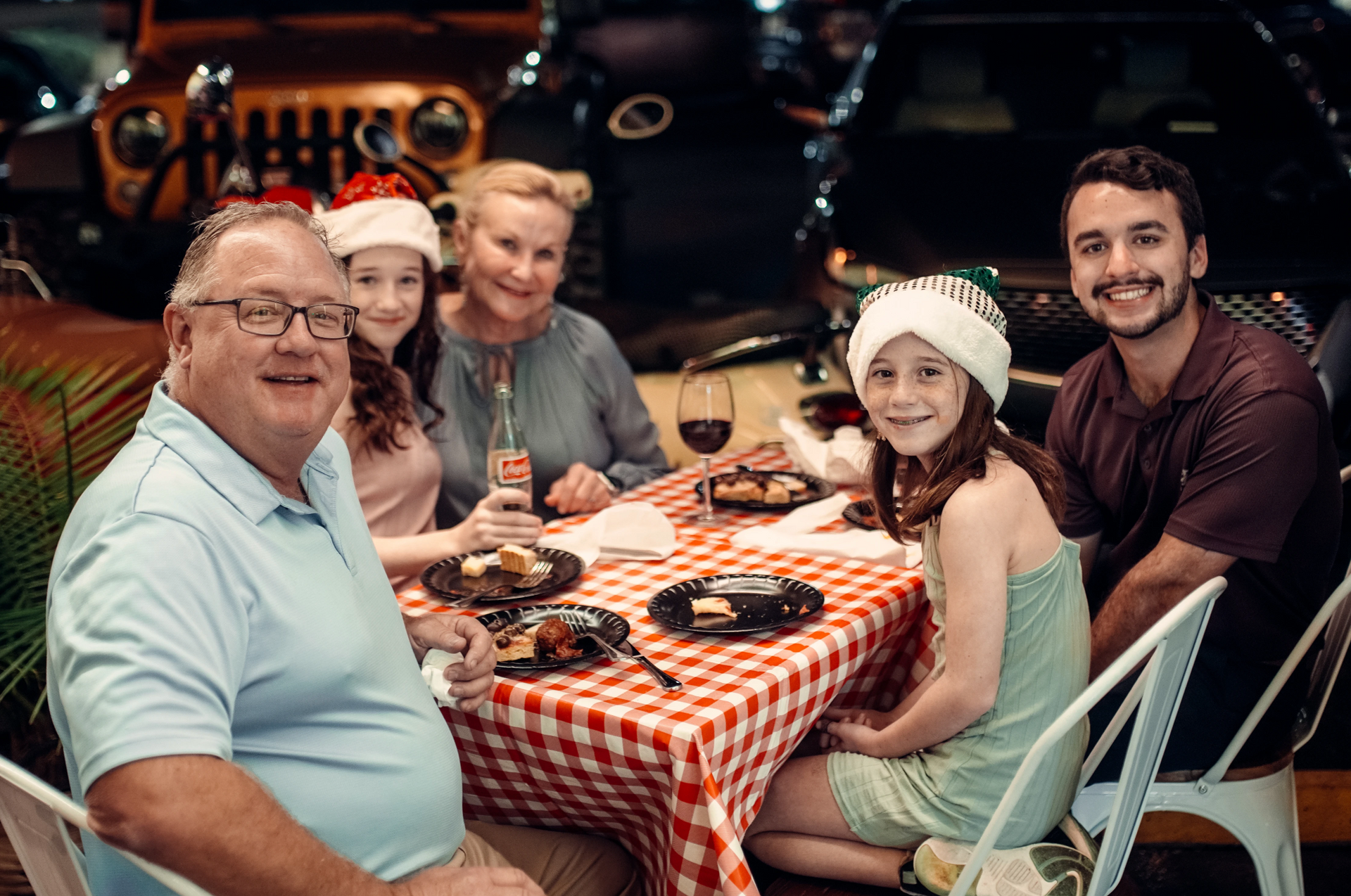 Family celebrating around a checkered tablecloth at a restaurant; people wear Santa hats, with cars in the background.