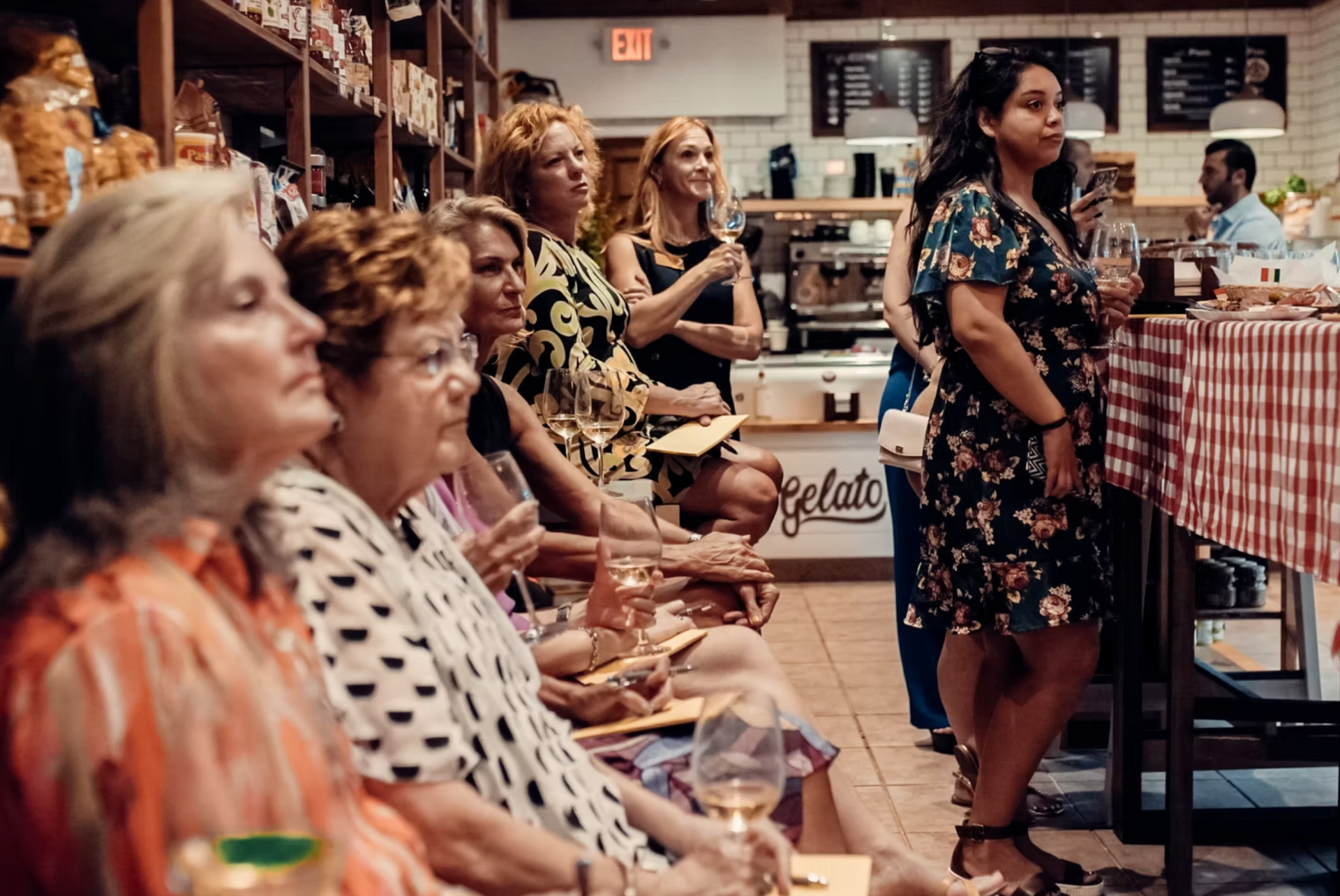 A group of women attending an event at a cafe with gelato signage, seated and standing, listening to a speaker.