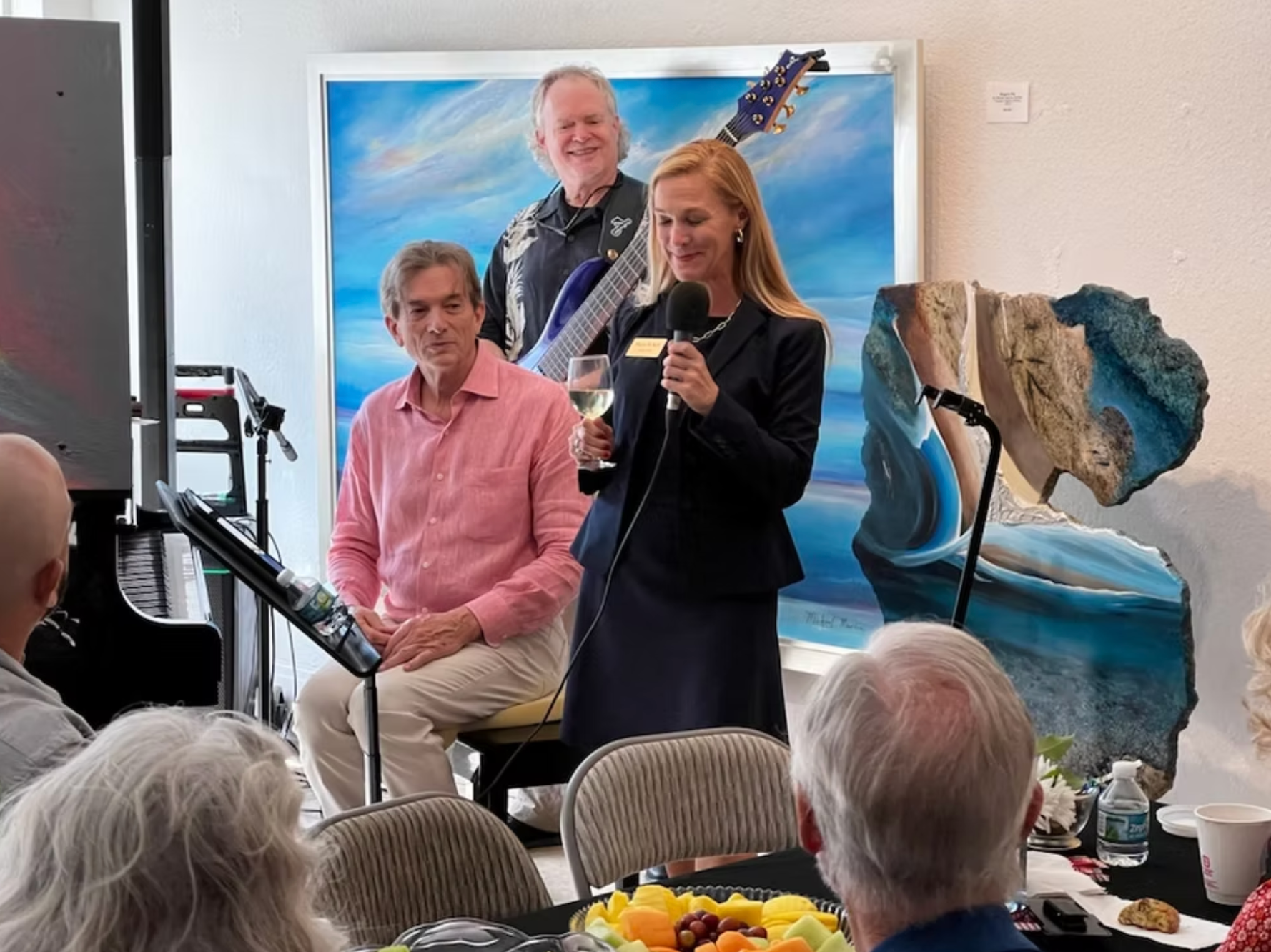 A woman in a suit speaks at a microphone, holding a glass, with two men and a band in the background.