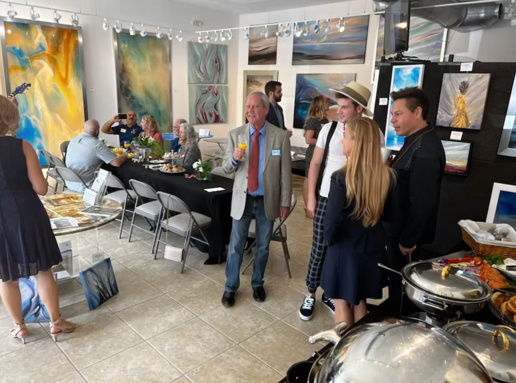 People at an art gallery opening, some holding drinks, with food on a buffet table and paintings on the walls.