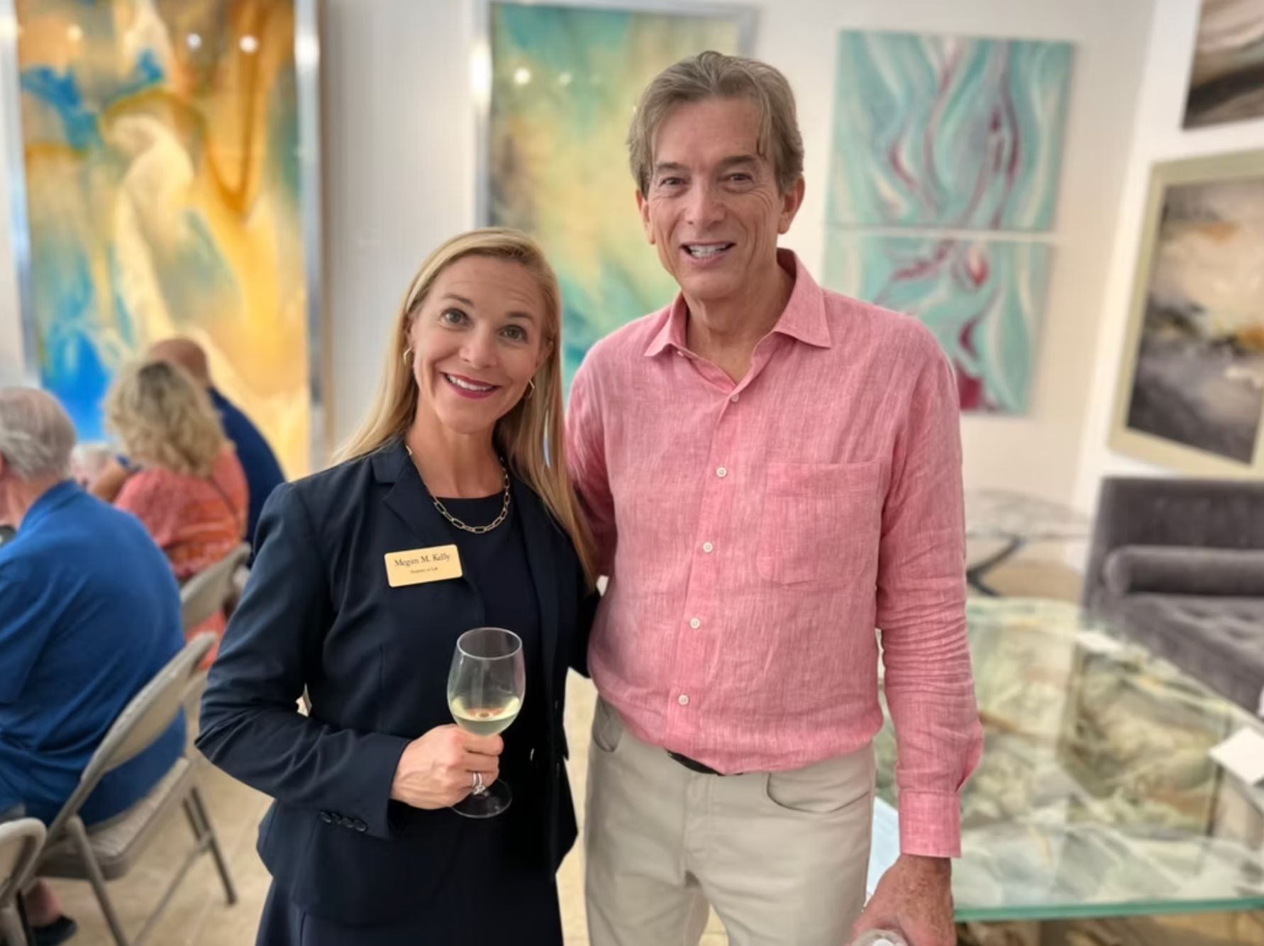 A smiling woman with a wine glass and a man pose together in an art gallery, surrounded by paintings.