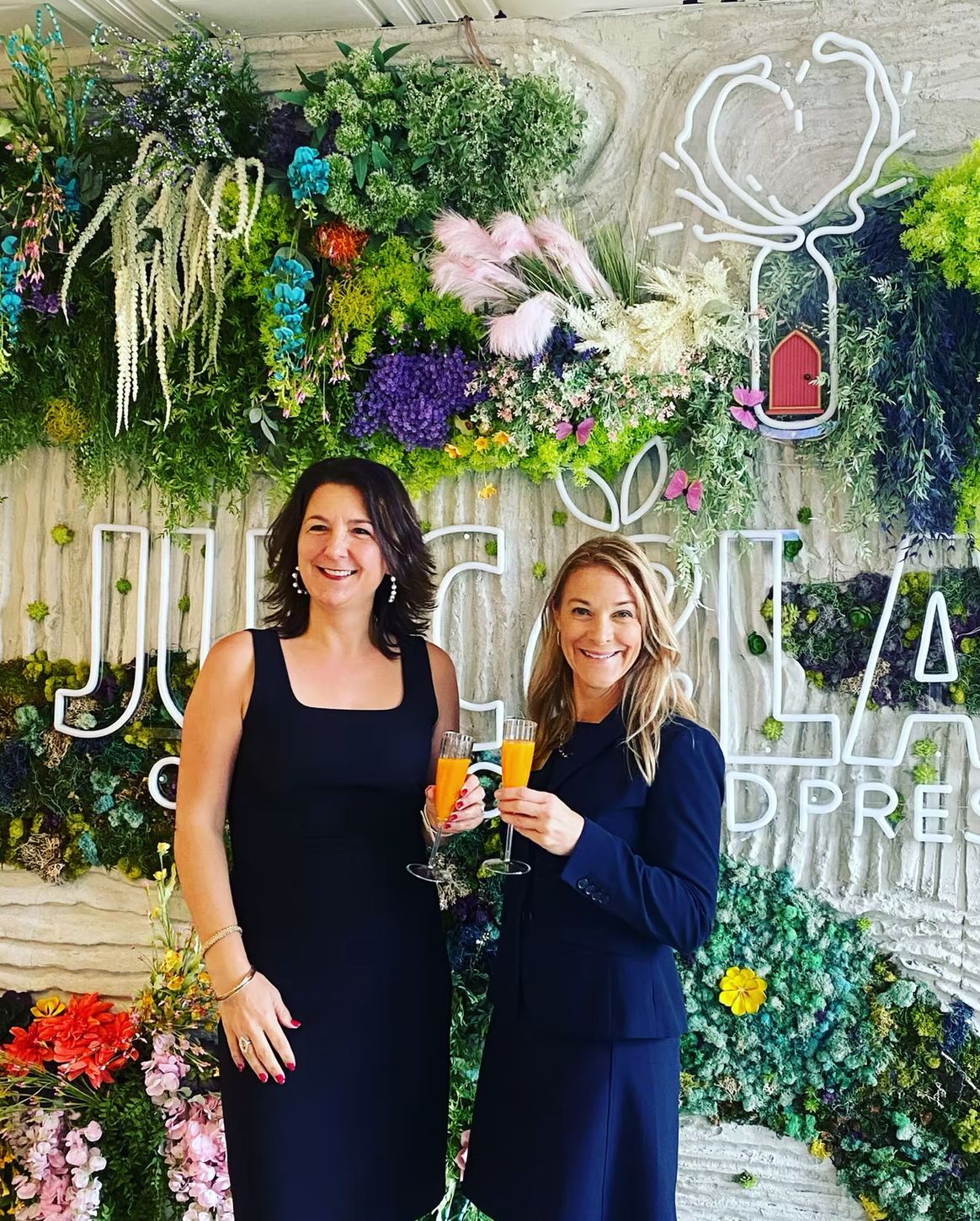 Two women in dresses holding drinks, smiling in front of a floral wall with neon signage.