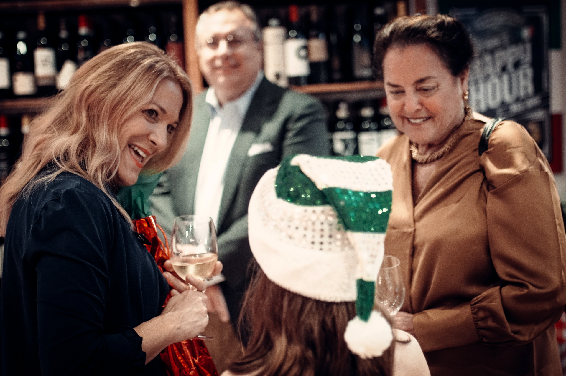 Three people smile and chat at a wine store, one wearing a Santa hat. Behind them, shelves of wine bottles.