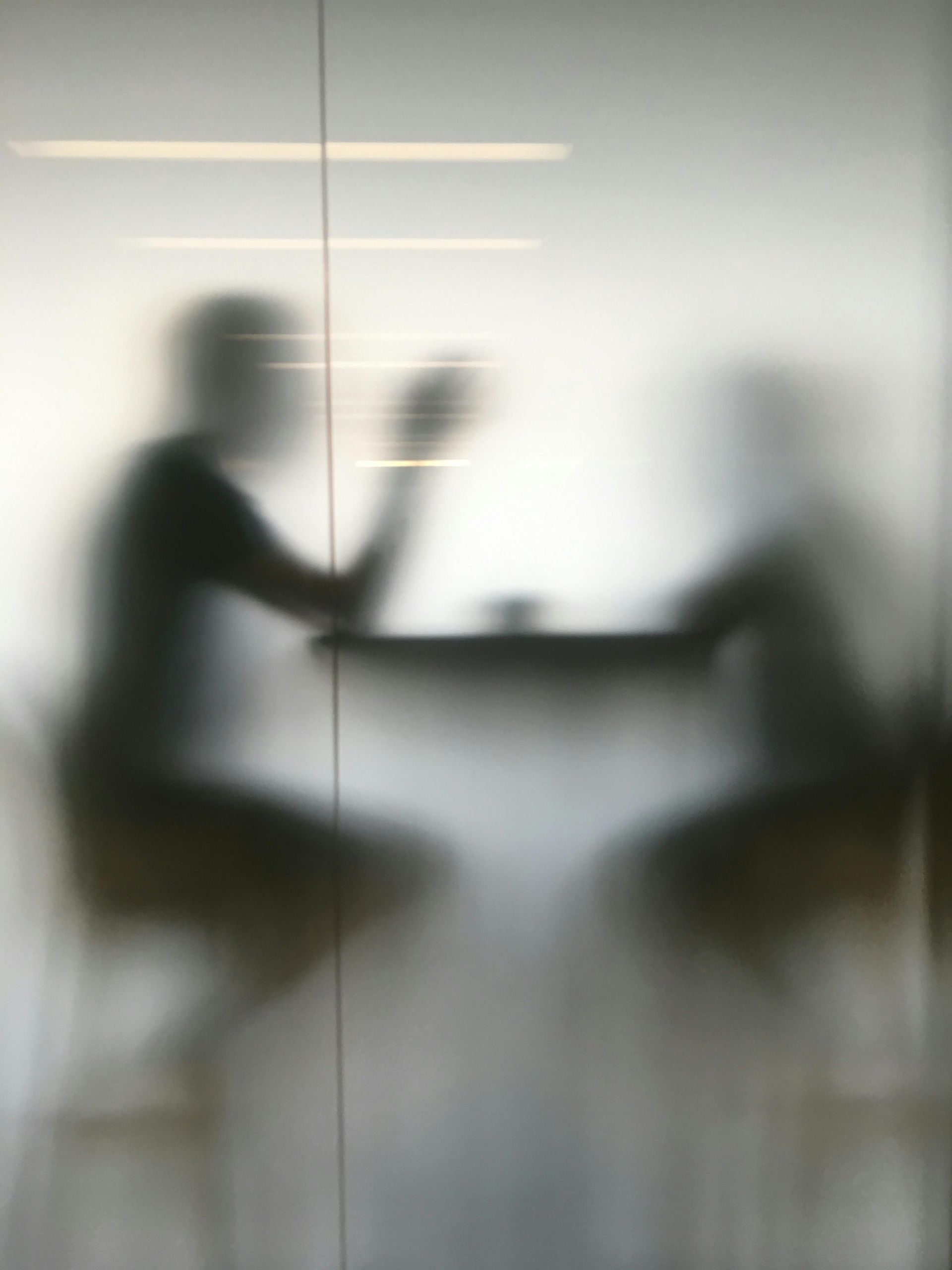 Two silhouetted figures seated at a table, behind frosted glass. One gestures with their hand, suggesting conversation.