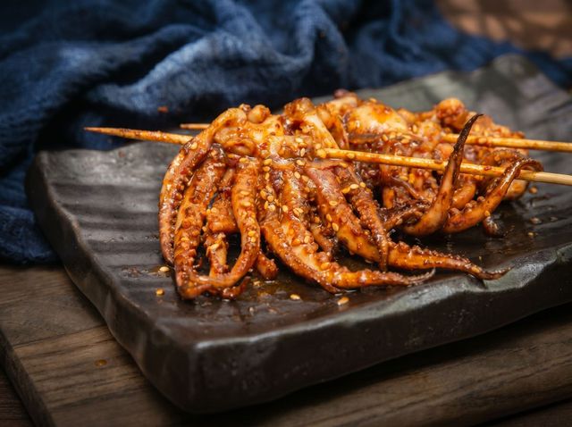 Spicy grilled squid skewers on a dark wooden plate, with sauce on a rustic table backdrop