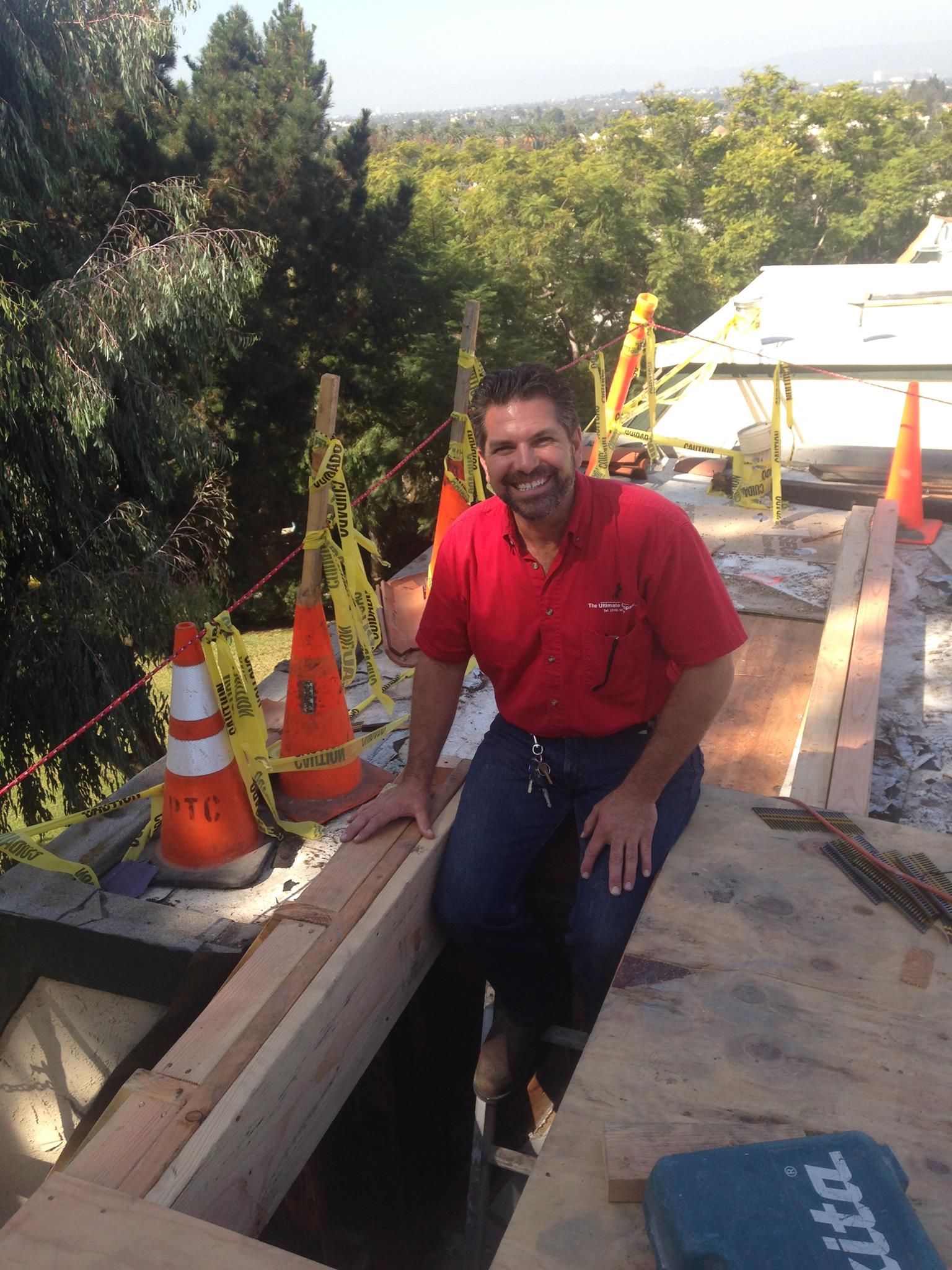 Man in red shirt and jeans smiles, sitting on a construction site, with trees in the background.