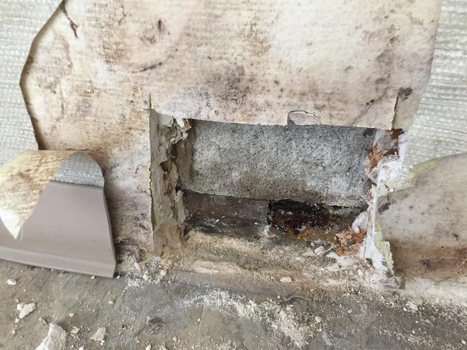 Hole in wall exposing brick interior, with peeling wallpaper and debris on concrete floor.