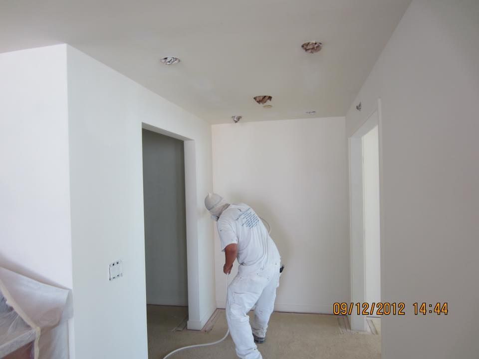 Person in protective suit painting white walls and ceiling of a room.