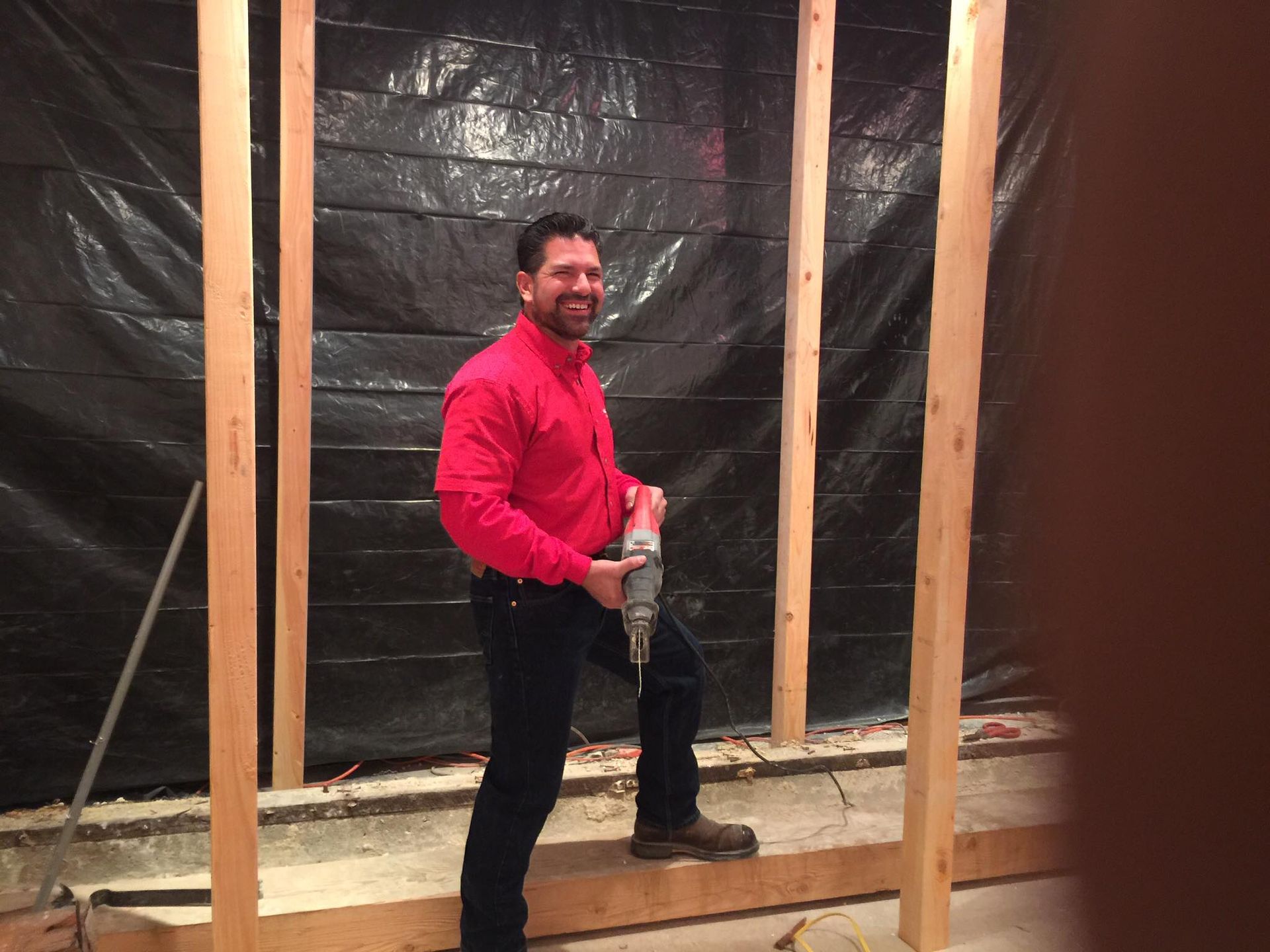 Man in red shirt holding a drill, standing in front of wooden studs, smiling. Black plastic background.