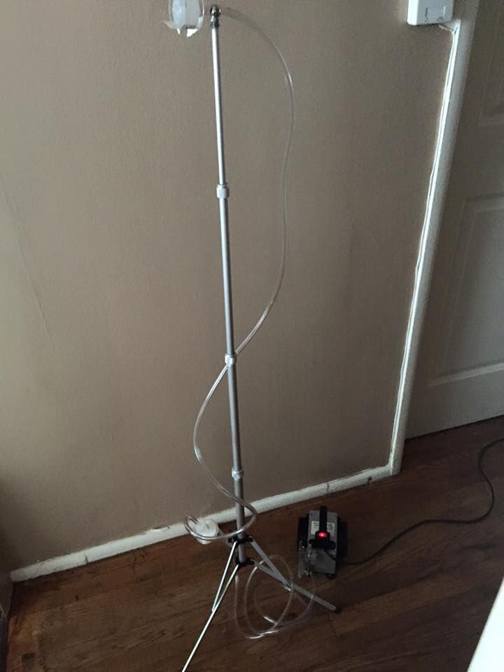 Metal IV stand with tubing and a foot-operated pump on a wooden floor near a wall.