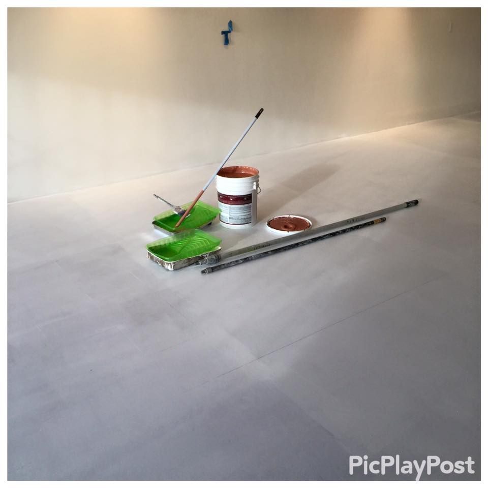 Paint supplies on a light gray floor, ready for painting a room. A paint can, trays, and roller extension are visible.
