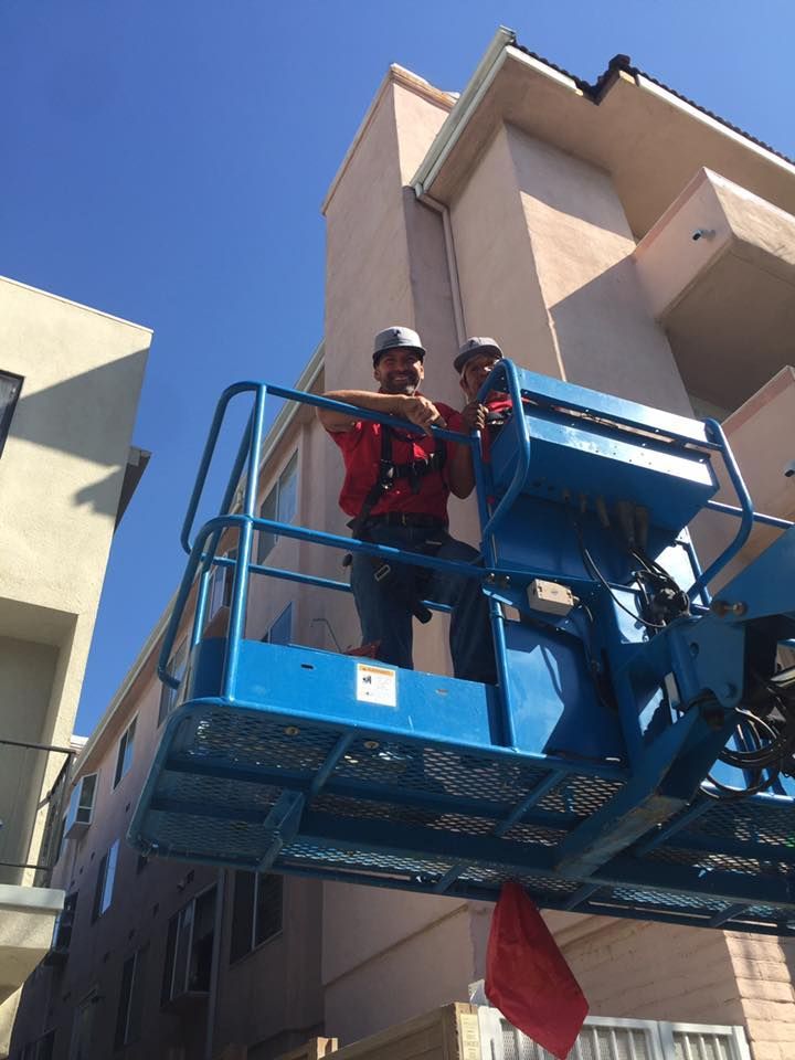 Two workers on a lift installing gutters on a multi-story beige building against a blue sky.
