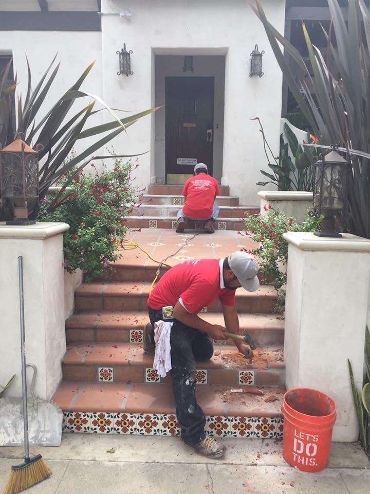 Two workers removing tiles from steps of a house. One kneels, the other sits, both wear red shirts.