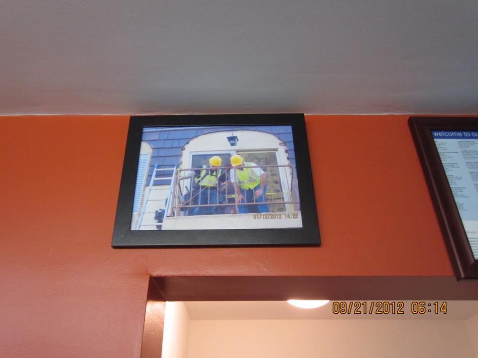 Two people in hard hats and vests on a balcony, framed photo on orange wall.