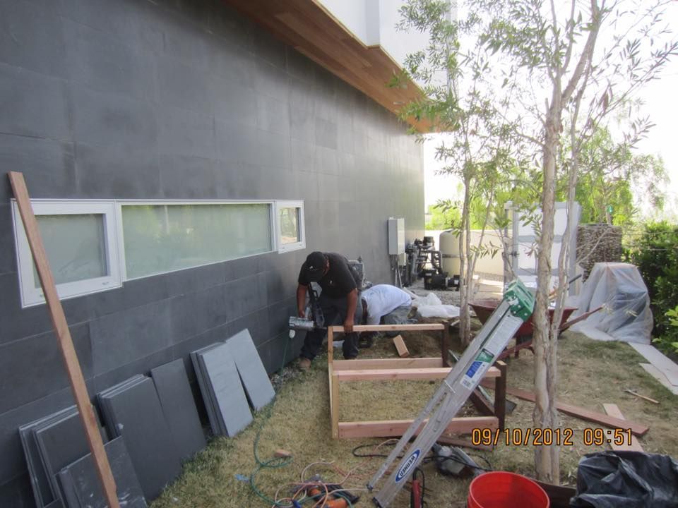 Construction worker cutting wood near a dark gray building; materials and tools scattered in the yard.