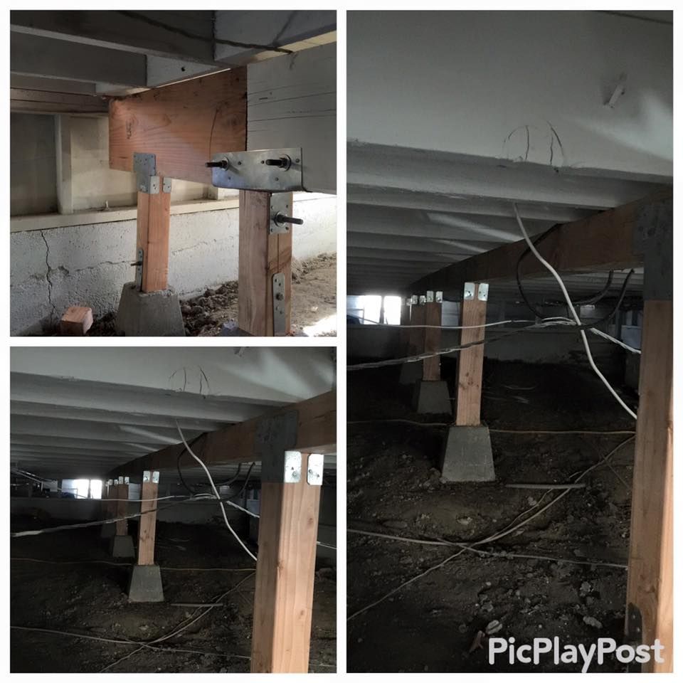 Wooden support beams under a building, some bolted to metal plates, concrete bases in a dirt crawl space.