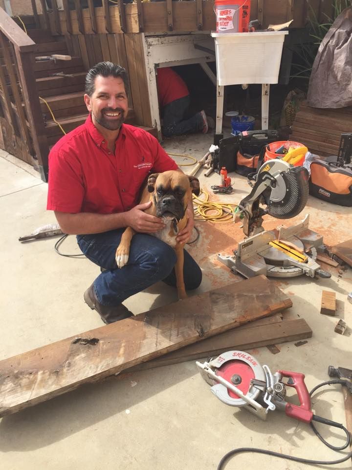 Man in red shirt, kneeling, holding dog, surrounded by woodworking tools on outdoor patio.