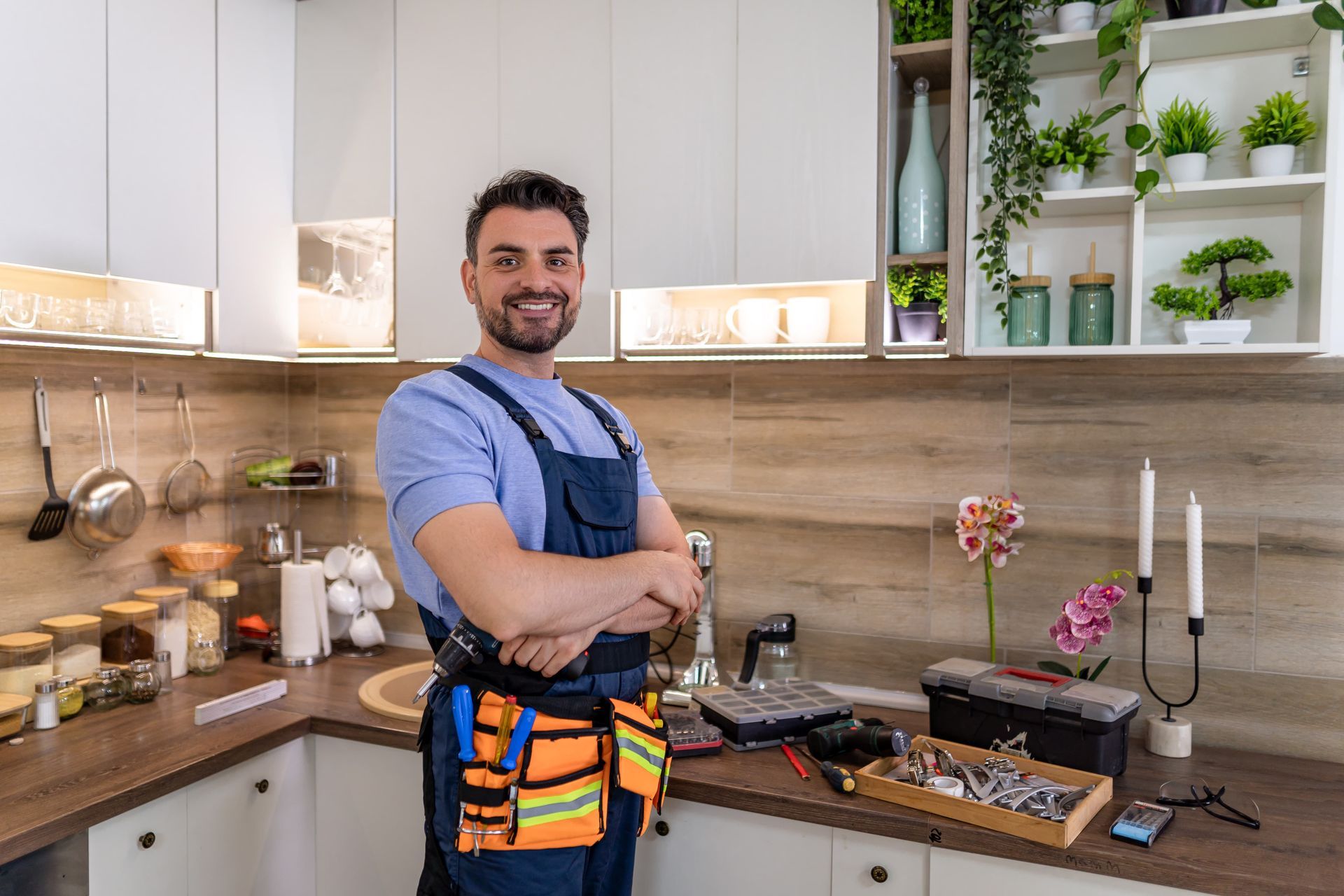 Plumber in overalls smiles, arms crossed, in a modern kitchen with tools on counter.
