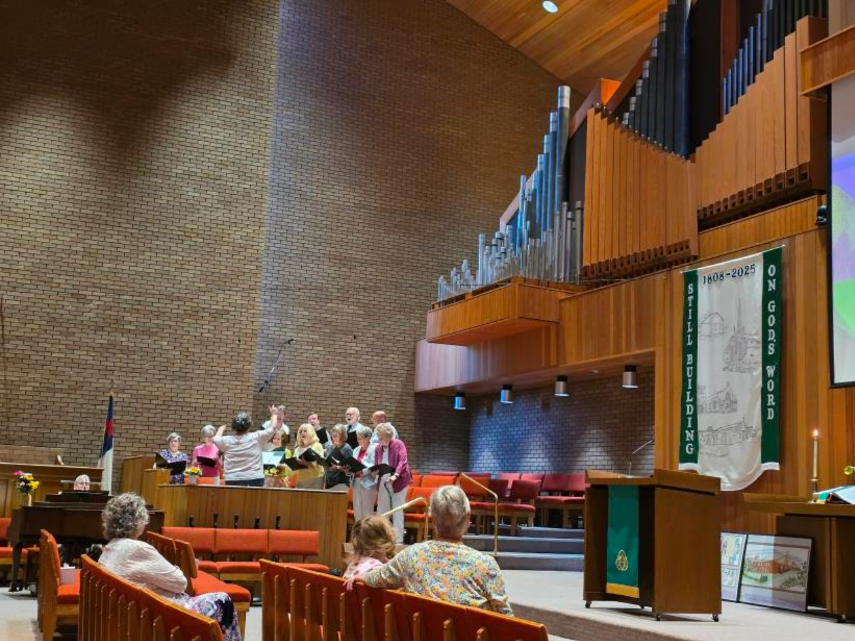Choir singing in a church. Brick wall, wooden pews, and organ pipes visible.