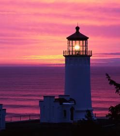 Lighthouse at sunset with bright light, in a vibrant pink and purple sky over the ocean.