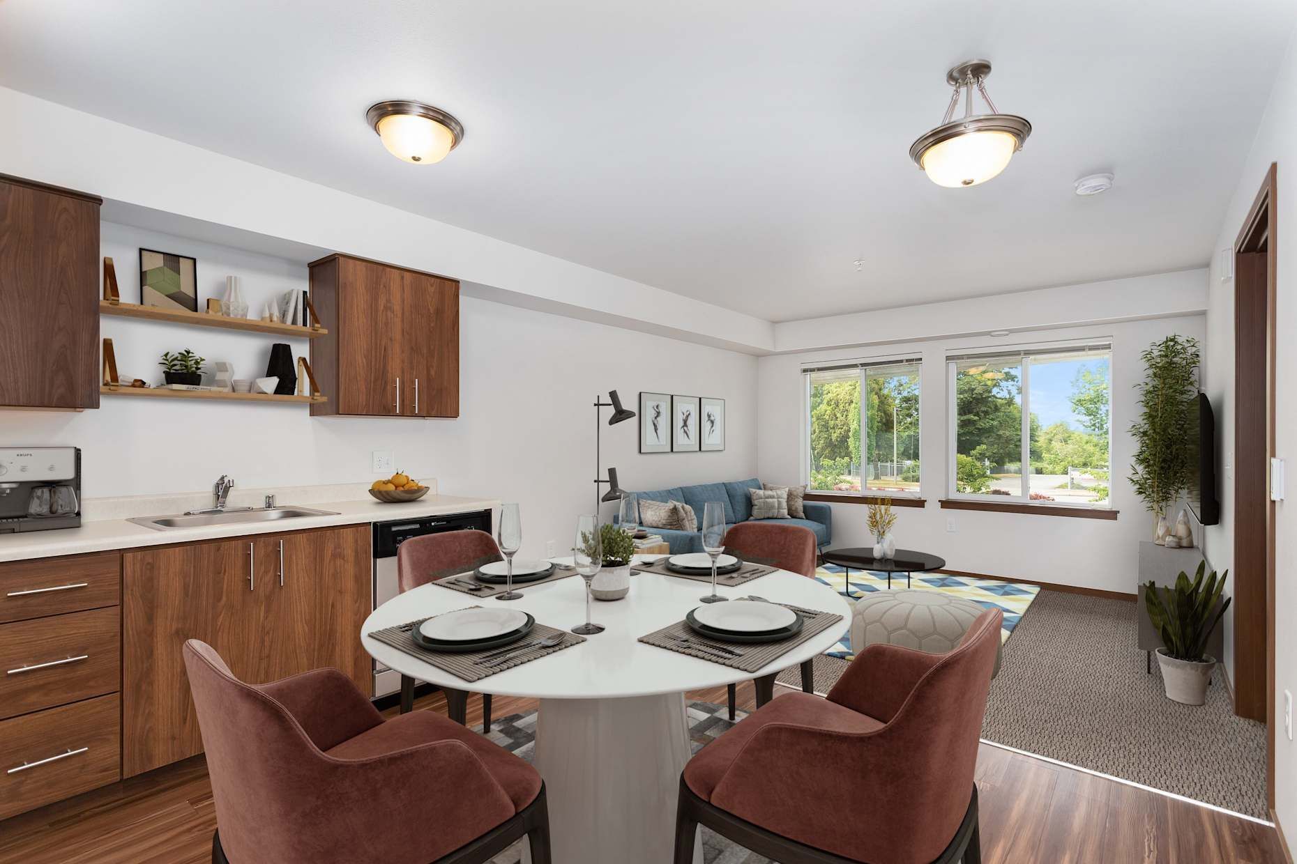 Open-concept living space: kitchen with wood cabinets, round dining table, living area with sofa, windows, and plants.