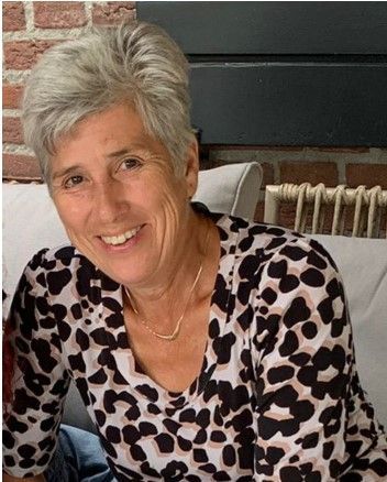 Een vrouw in een roze jasje staat voor een bakstenen gebouw.