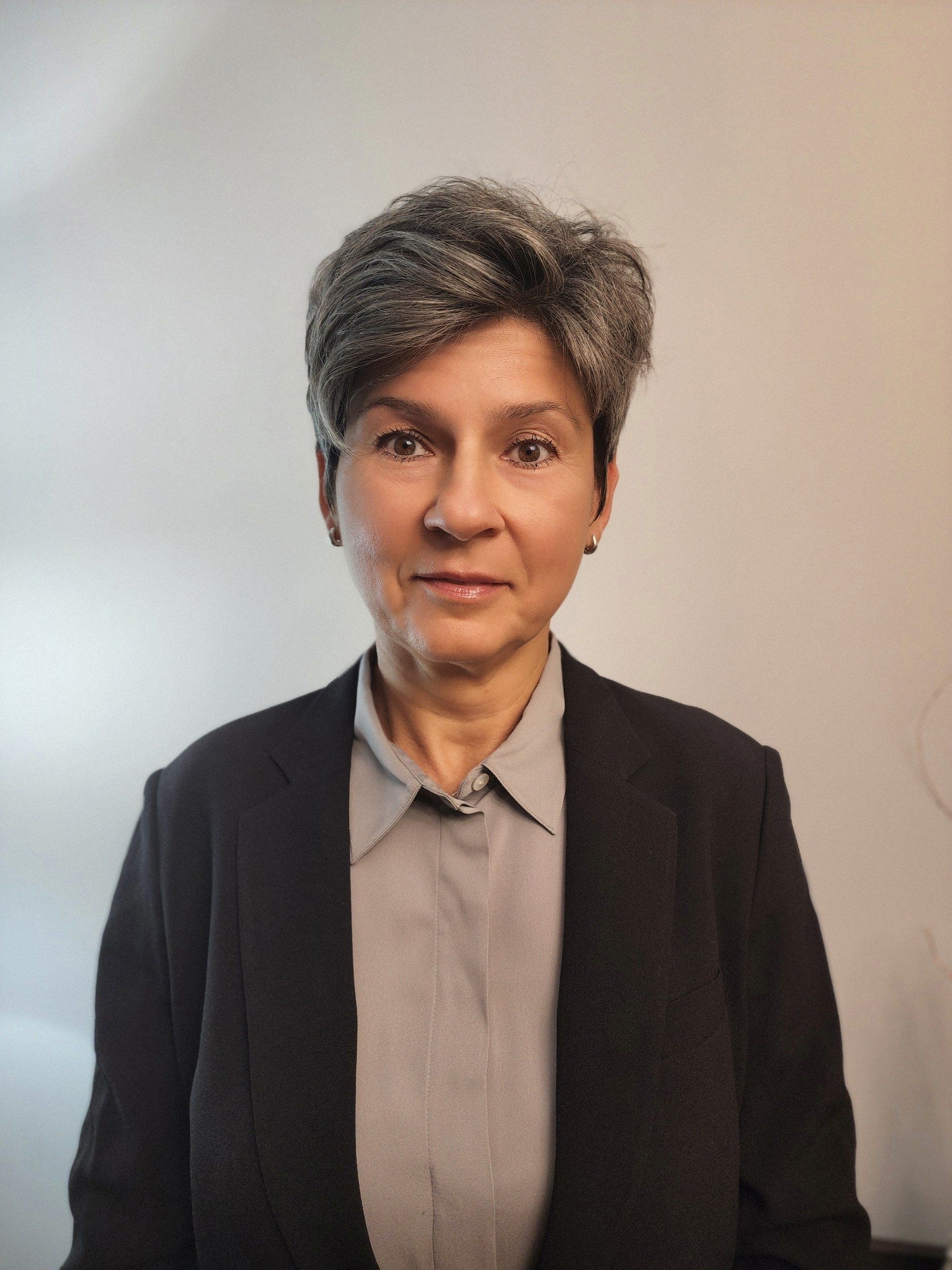 A woman wearing a black jacket and a grey shirt is standing in front of a white wall.