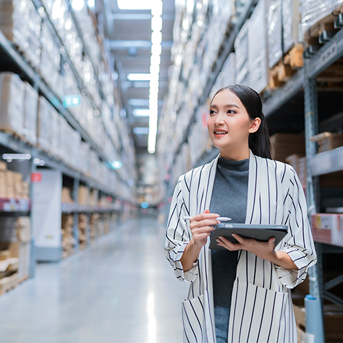 A woman is standing in a warehouse holding a tablet and a pen.