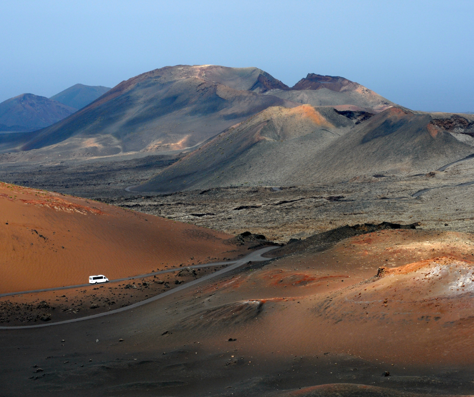 A car is driving down a dirt road in the desert with mountains in the background.