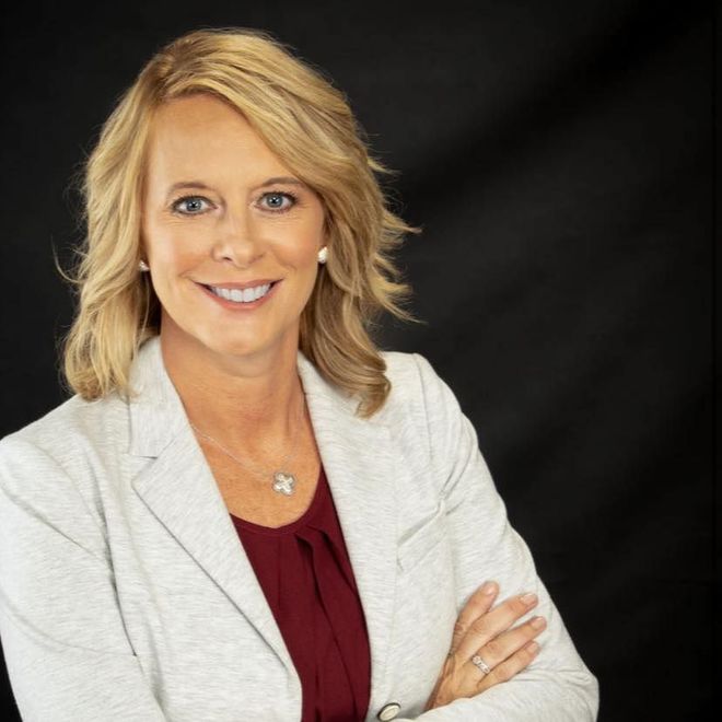 Blonde woman with arms crossed smiling, wearing a gray blazer over a maroon shirt, against a black backdrop.