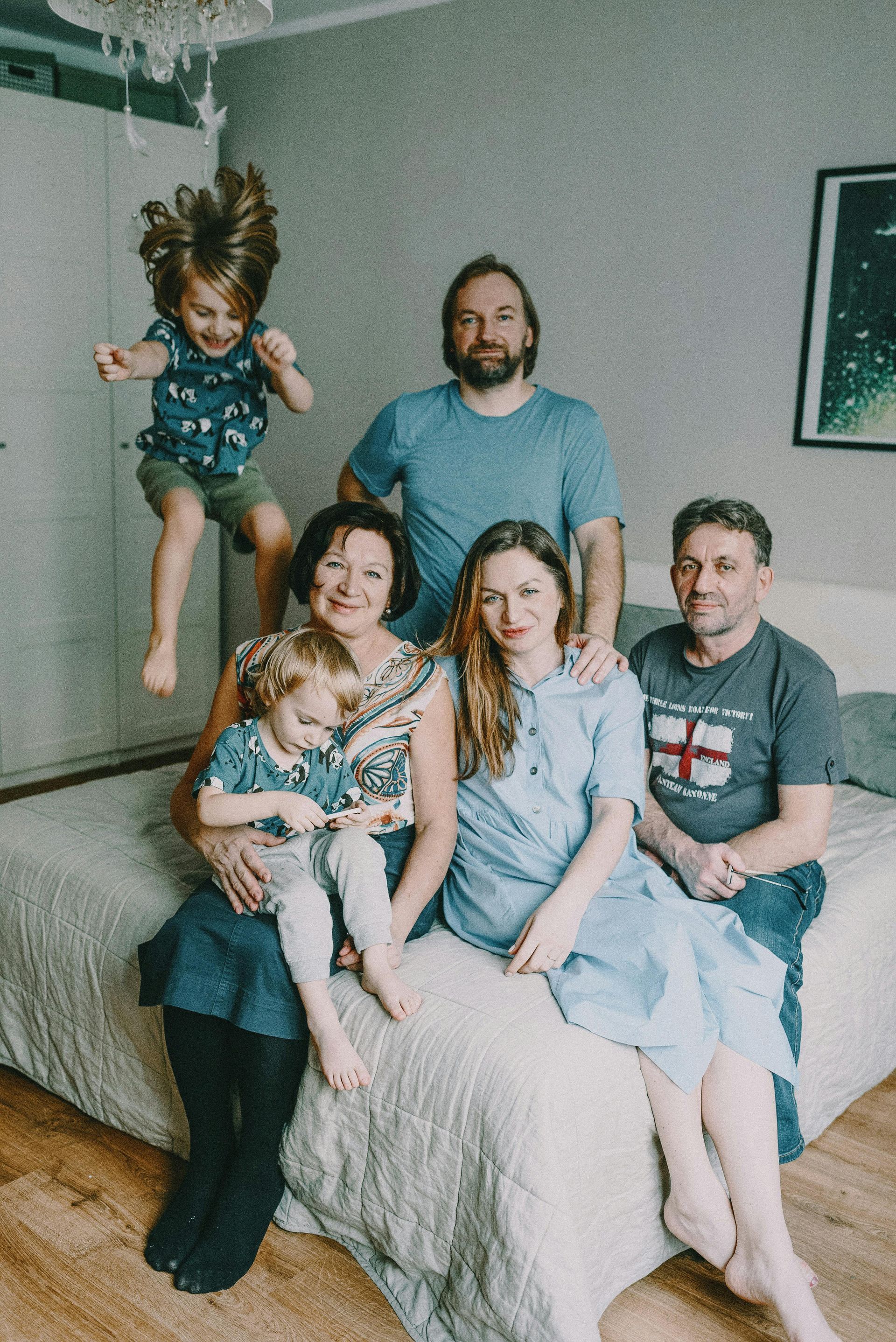 Family portrait with a child jumping mid-air. Six people, including children and adults, are on a bed in a bedroom.