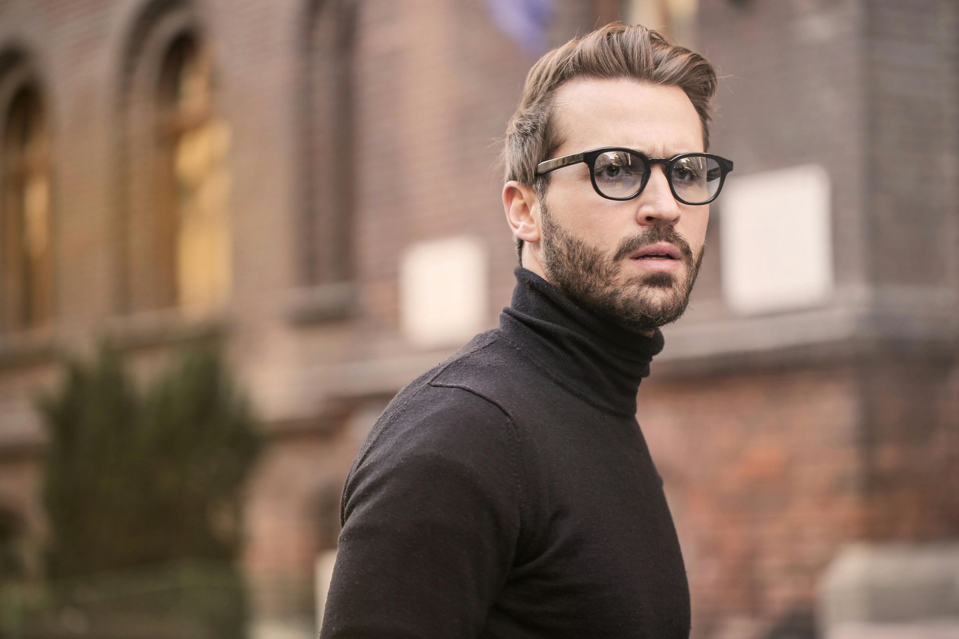 Man with beard and glasses in a black turtleneck, looking slightly surprised, outdoors near a building.