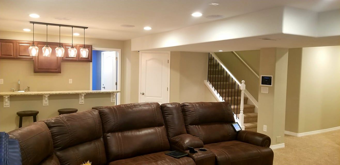 A basement living space featuring a brown leather sectional, a wet bar with stools and pendant lighting, and a staircase.