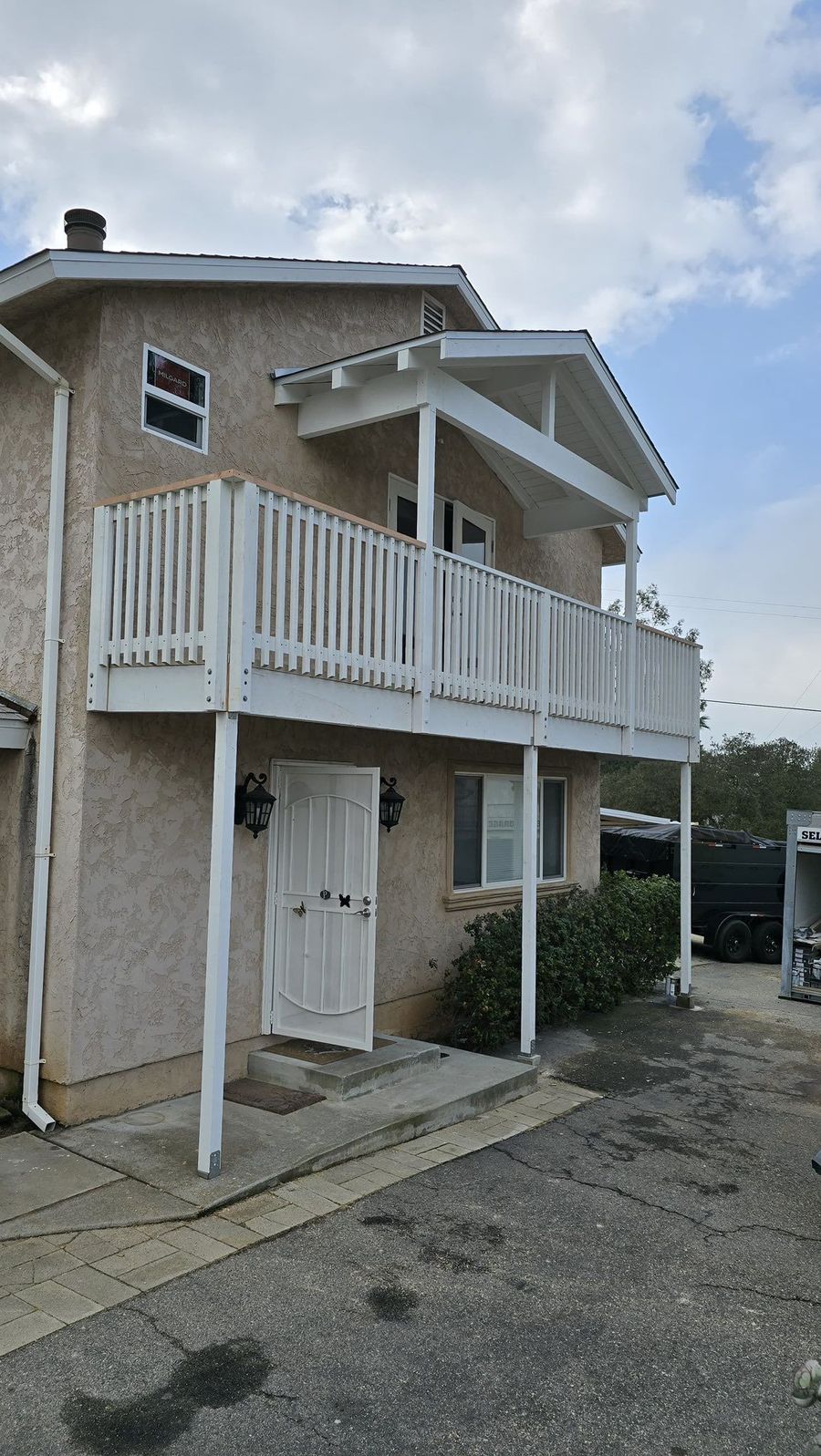 A two-story, beige stucco house with a white wooden balcony and a white front door, set against a cloudy blue sky.