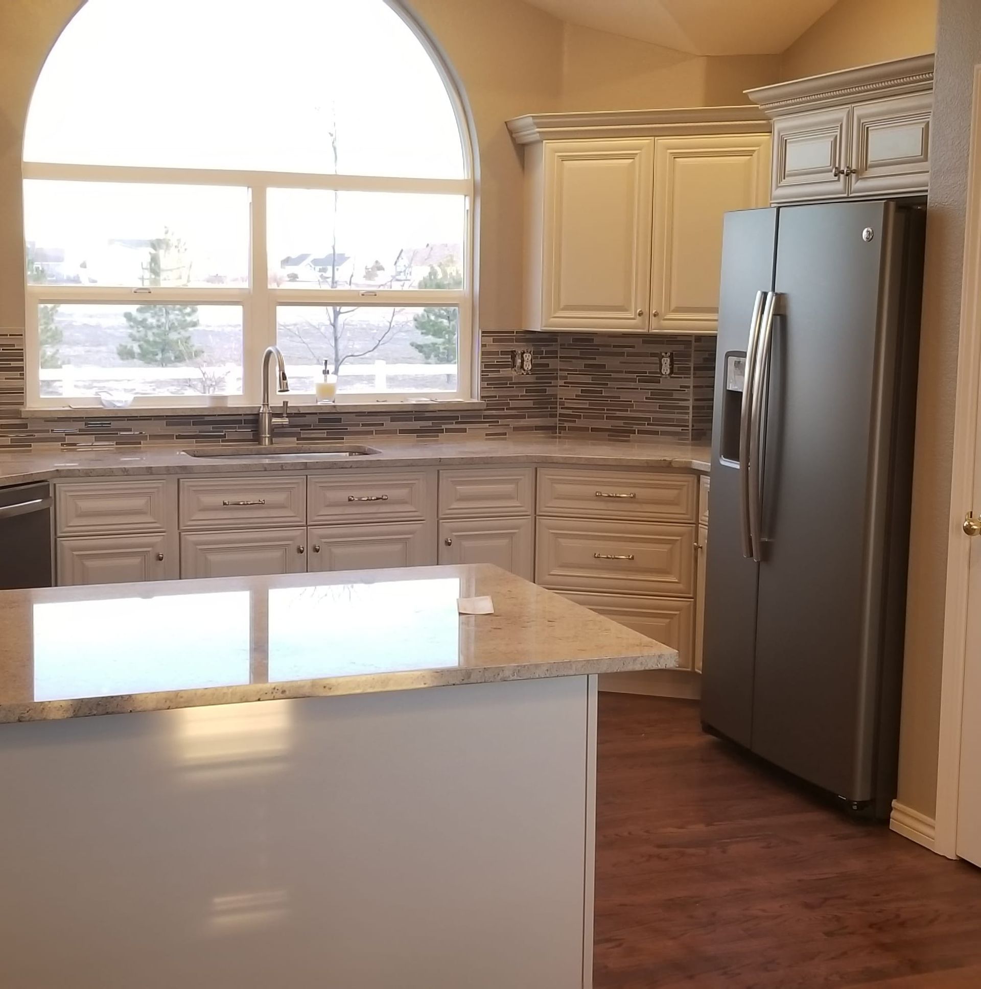 A modern kitchen featuring off-white cabinets, granite countertops, a stainless steel refrigerator, and a large window.