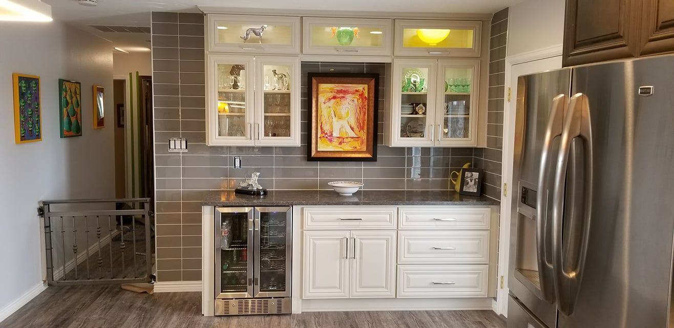 A white kitchen cabinet unit with glass doors, a wine cooler, and a framed painting against a grey tiled backsplash.
