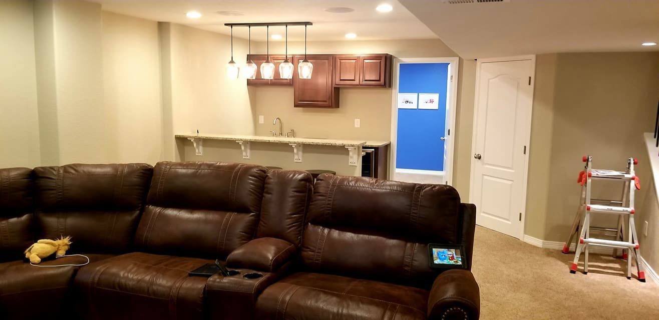 A brown sectional sofa sits in front of a basement bar area with wooden cabinets, a sink, and a step ladder nearby.