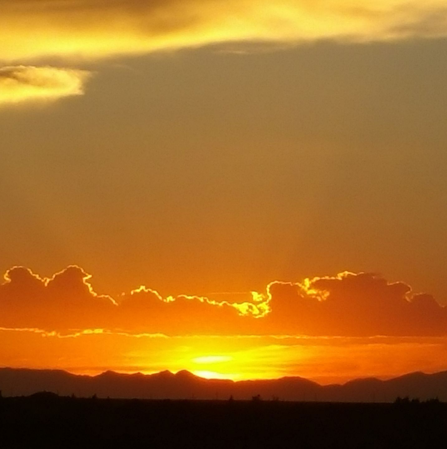 A vibrant orange and gold sunset illuminates clouds above a dark, silhouetted mountain range.
