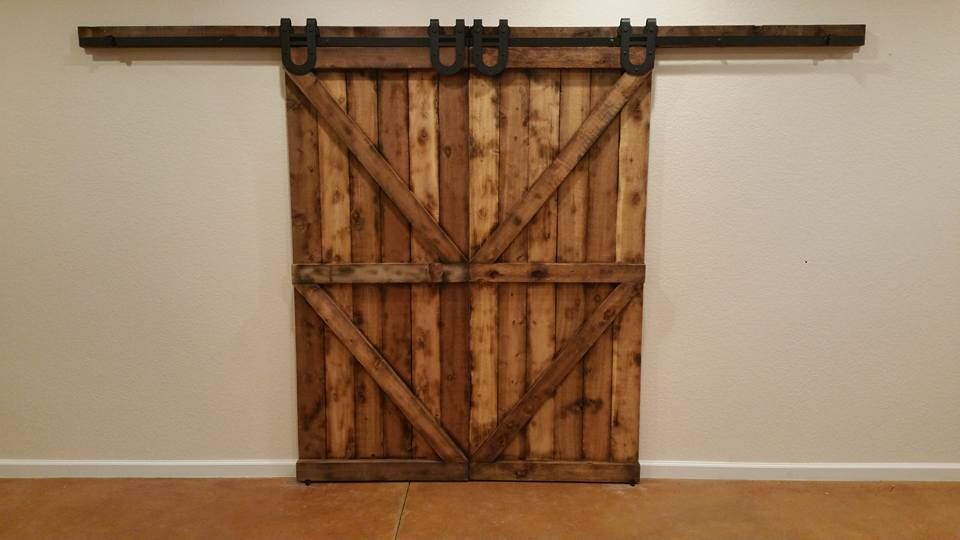 Double rustic wooden barn doors with X-bracing, hung on a dark metal track against a plain white wall.