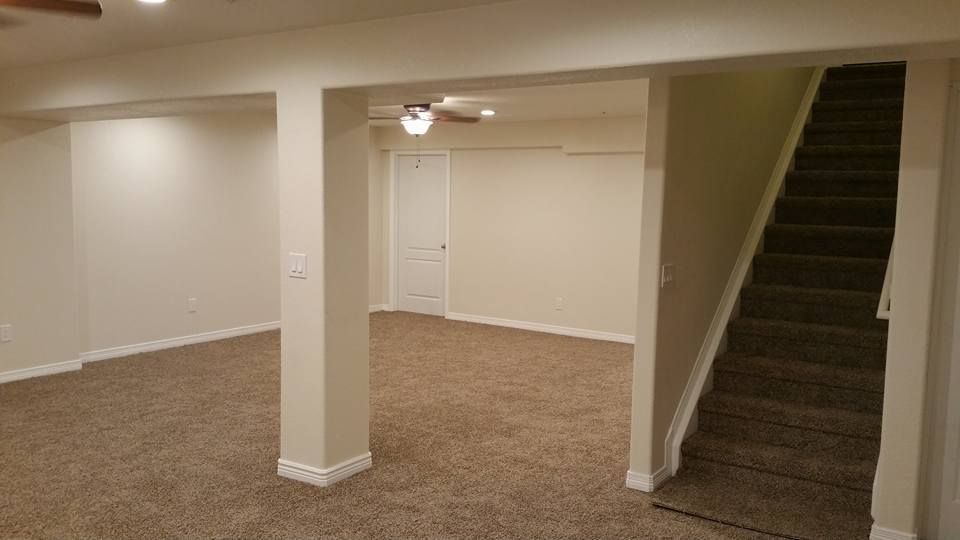An empty, beige-carpeted basement room with a support pillar, a white door, and stairs leading upward.