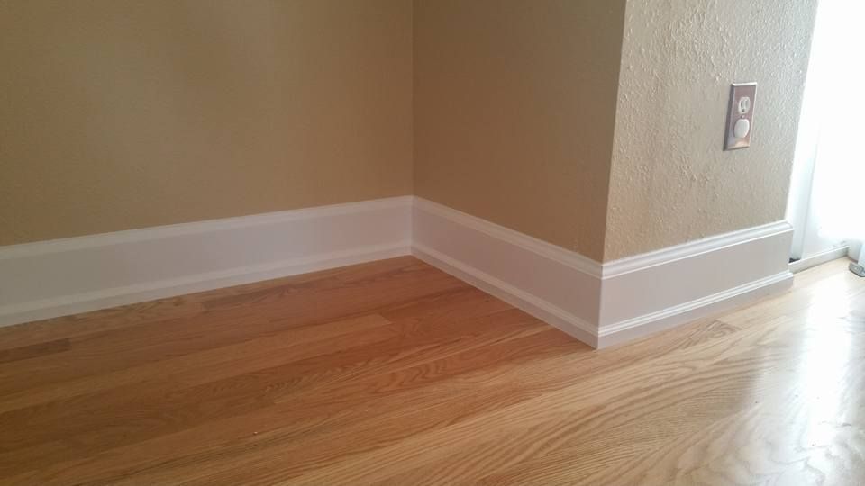 White baseboards installed against a corner of beige walls and hardwood flooring.