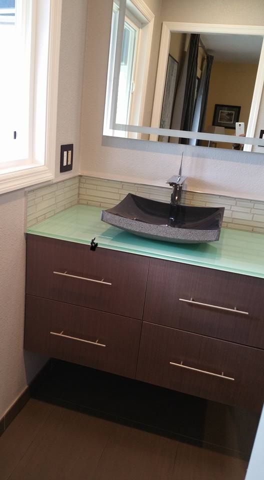 A modern bathroom vanity featuring a dark rectangular vessel sink, light green glass countertop, and brown cabinetry.