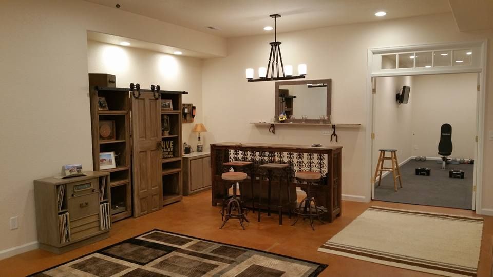 A home basement features a bar with stools, a wood storage unit, and a doorway leading to a small gym area.