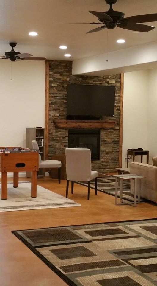 A furnished living room with a stone fireplace, television, two ceiling fans, and a foosball table on a concrete floor.