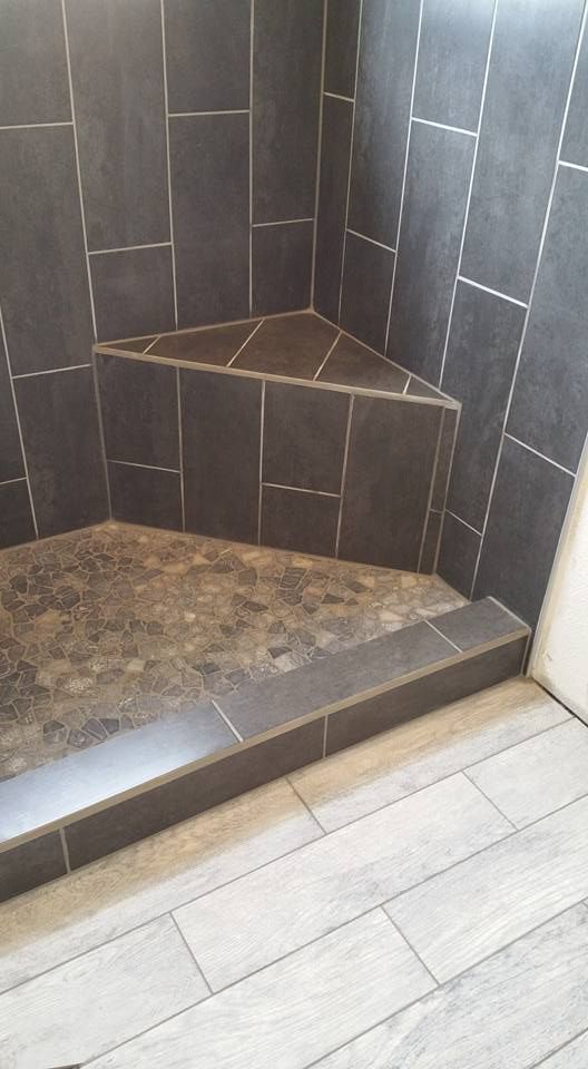 A shower stall with dark gray wall tiles, a built-in corner seat, and a pebble-tiled floor, separated by a tiled curb.