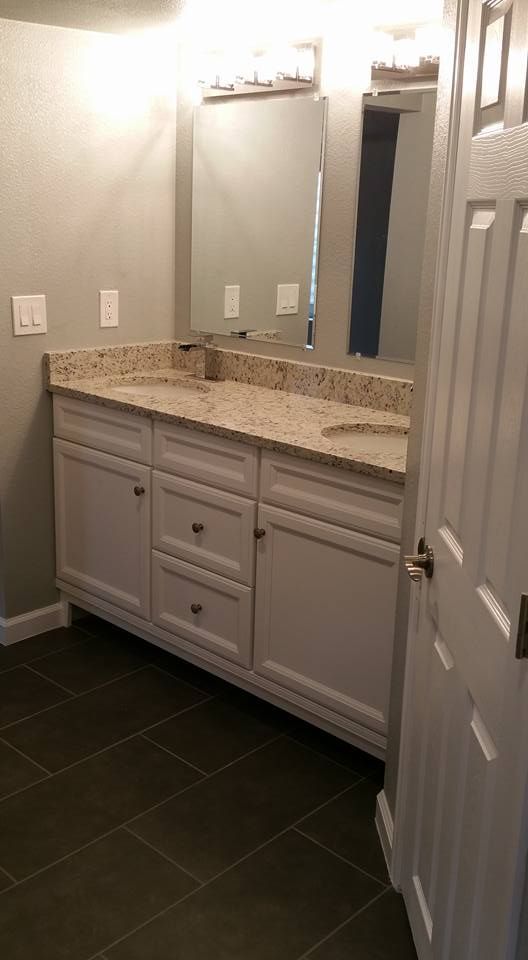 A bathroom vanity with a speckled granite countertop, white cabinets, dual sinks, and mirrors on a grey tiled floor.