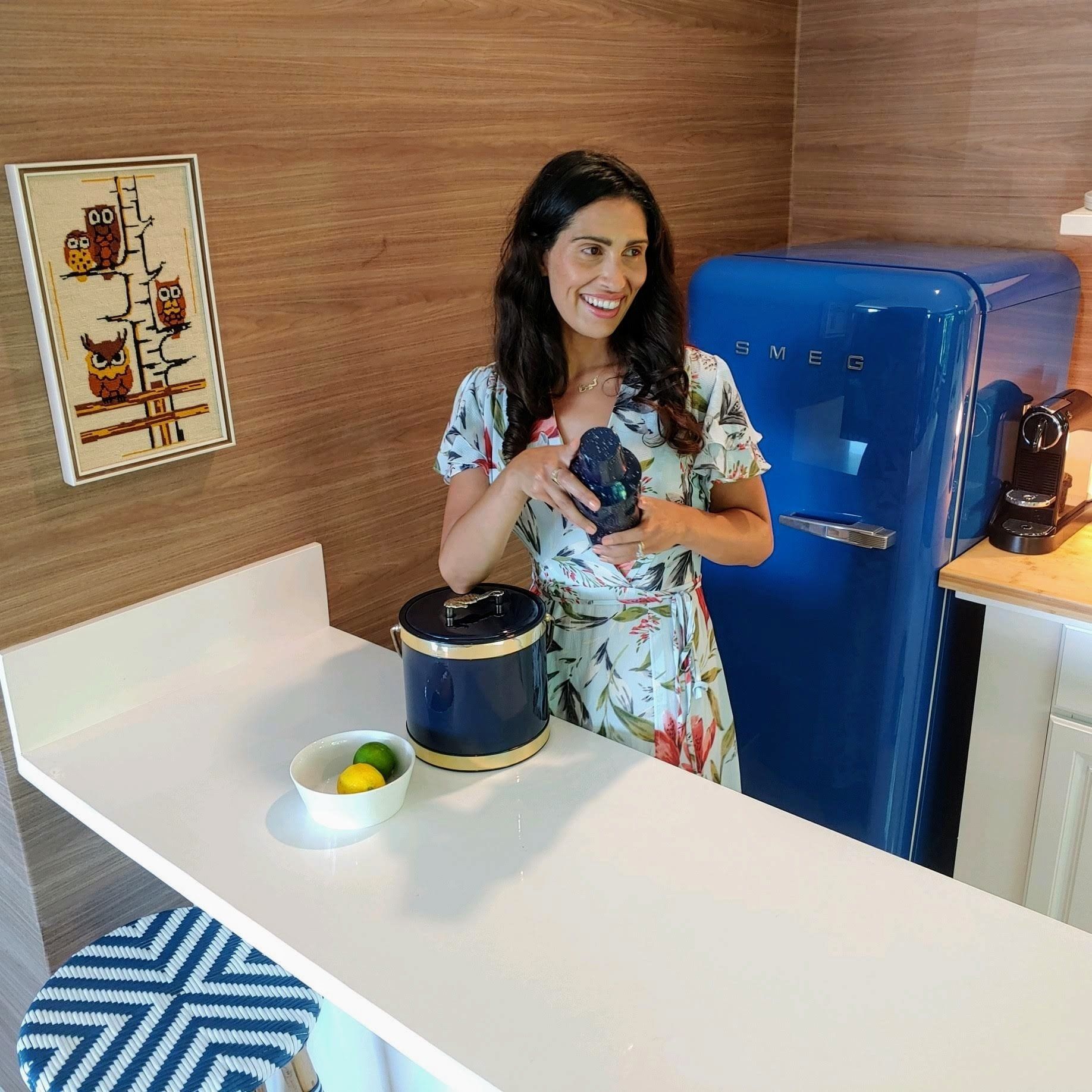 Woman smiles in kitchen with blue fridge, holding object. Wooden wall, white counter, owl art.