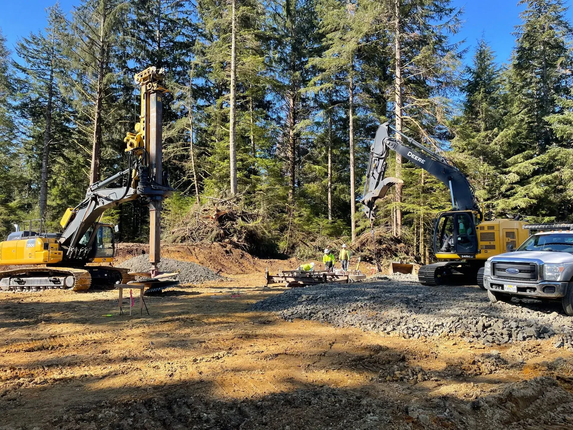 Two excavators working on a construction site surrounded by trees; sunny day.
